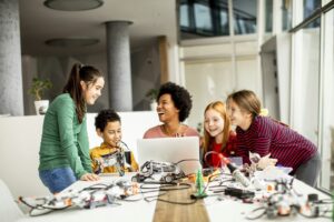 Docente y estudiantes observan materiales científicos, una computadora y componentes de robótica sobre una mesa, escena vinculada con la Licenciatura en Bioinformática a distancia.
