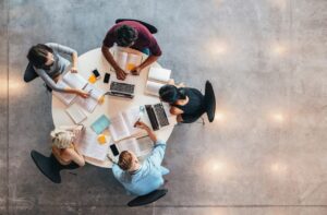 Cinco estudiantes trabajan alrededor de una mesa con computadoras portátiles, cuadernos, hojas y marcadores, escena de estudio de la Licenciatura en Enfermería.